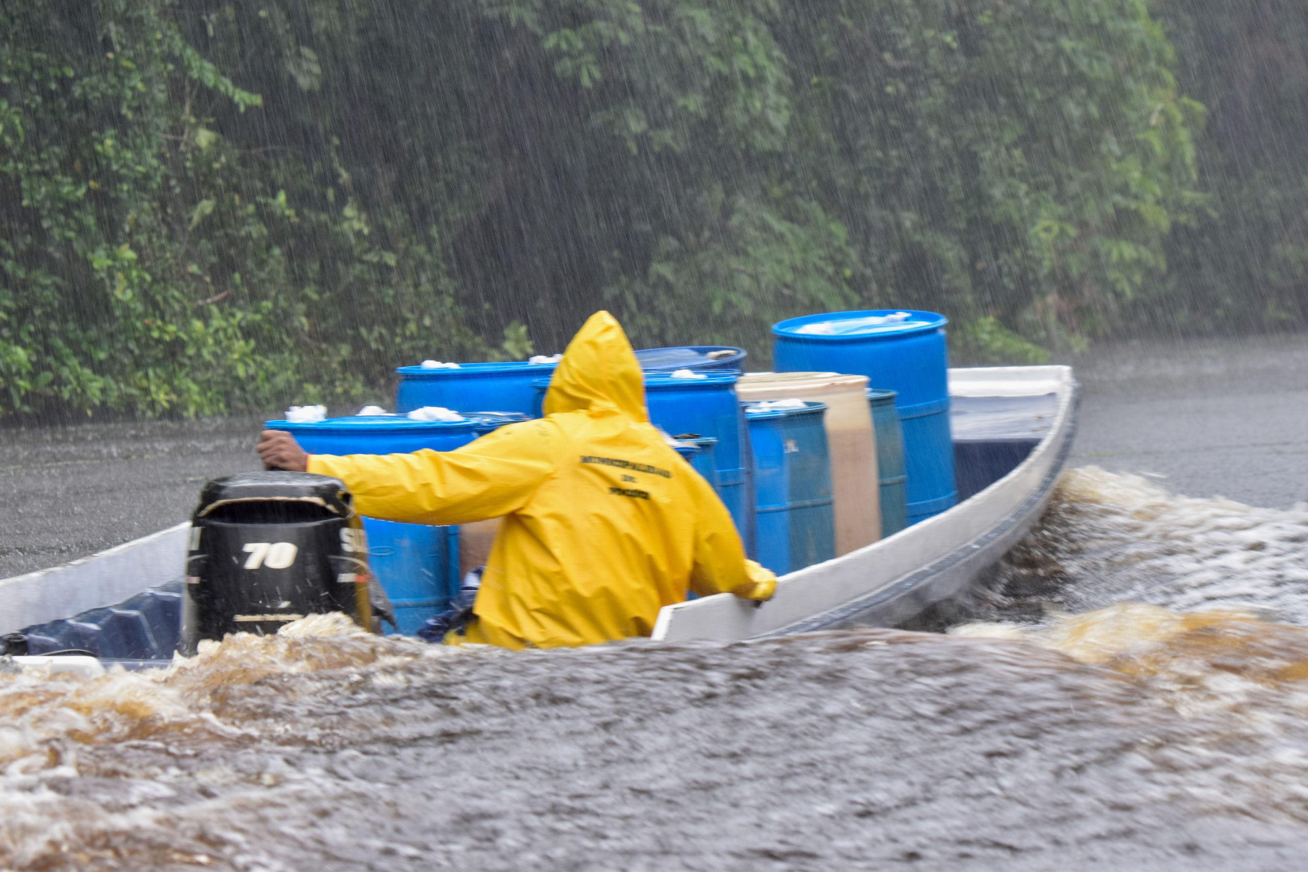 Small white boat on the river with man in yellow raincoat steering it through the rain.  Sitting very low in the water. Boat is transporting drums of water