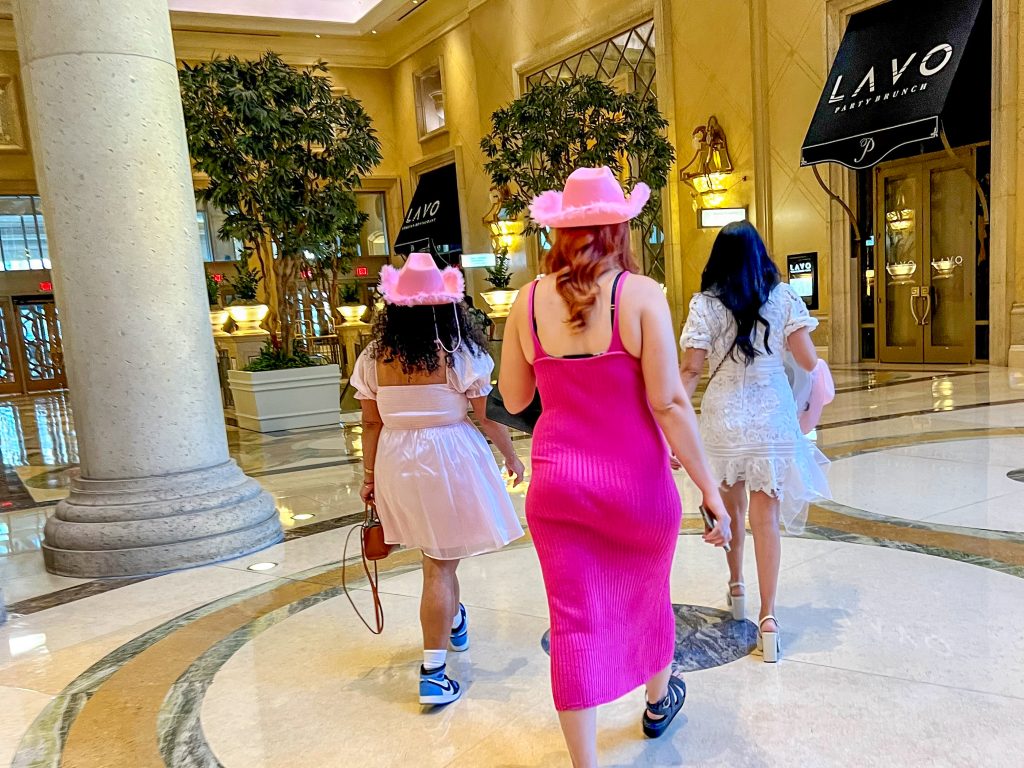 Three ladies walking away from the camera towards the door of a hotel lobby.  The one closest to the camera is wearing a bright pink dress and pink cowboy hat, the lady on the left has a pale pink dress and cowboy had and the lady on the right has a white dress but no hat