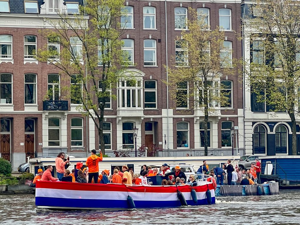 Tub boat covered in Dutch flag on the canal with party goers on board. A second party boat is behind it. Dutch houses in the background on the other side of the canal